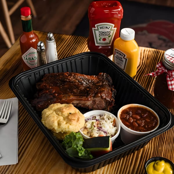 Ludy's BBQ ribs, biscuit, coleslaw, and baked beans in a to-go container with condiments.