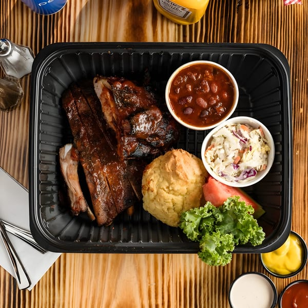 Ludy's BBQ ribs meal with baked beans, coleslaw, cornbread, and watermelon in a black container.