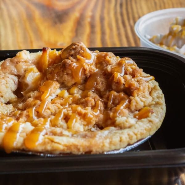 Ludy's BBQ peach cobbler with crumble topping and caramel drizzle in a black container.