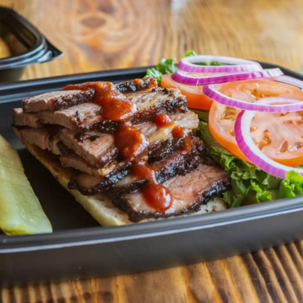 Ludy's BBQ brisket sandwich with sauce, lettuce, tomato, red onion, and a pickle spear.