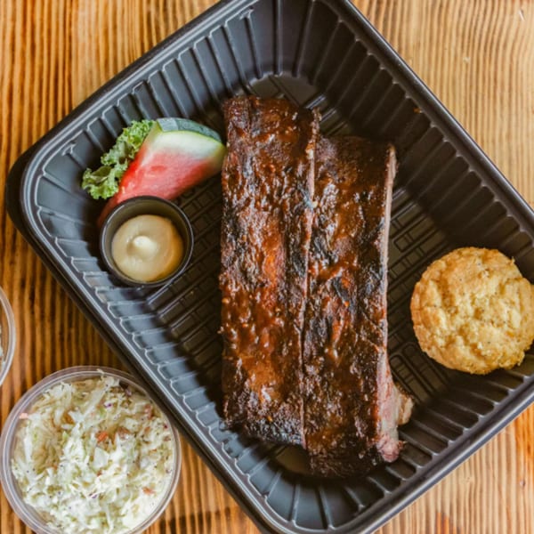 Ludy's BBQ ribs, cornbread, coleslaw, watermelon, and sauce in a black to-go container.